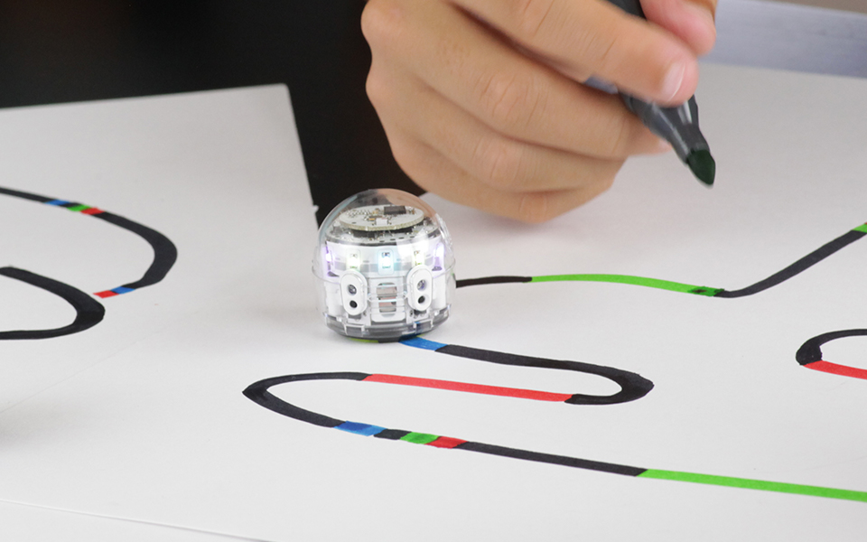 A small coding bot known as an ozobot sits on a white paper with black, red, blue, and green paths on it.