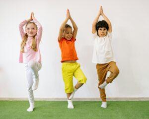 Three children doing a yoga pose with their hands above their head while balancing on one leg.