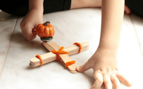 Child-sized hands play with a handmade catapult made of rubber bands and popsicle stickers. An orange, plastic pumpkin sits in the launcher.